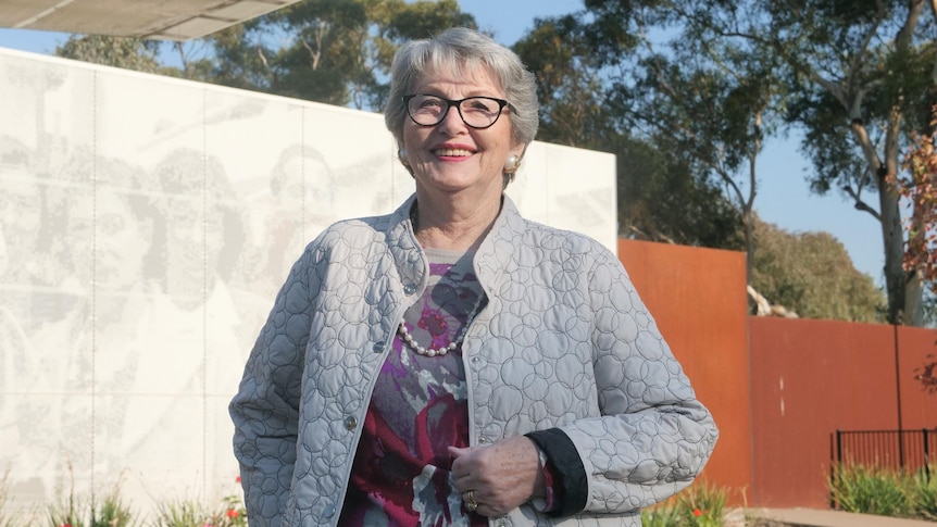 An elderly woman wearing glasses and a grey coat and pearls is standing outside. 