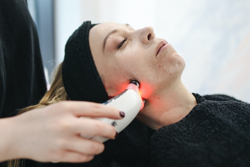 A woman lies on a salon bed as a laser tool is used on her face