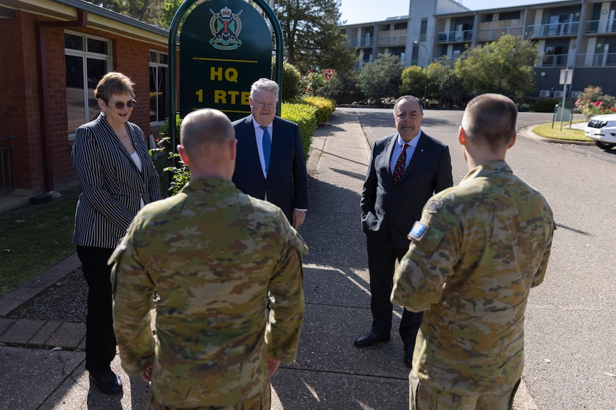 A group of three people in civilian clothes talk with army recruits in uniform