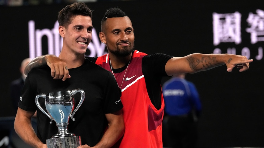 Nick Kyrgios points and puts his arm around doubles partner Thanasi Kokkinakis, who is holding the Australian Open trophy.