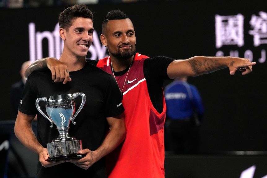 Nick Kyrgios points and puts his arm around doubles partner Thanasi Kokkinakis, who is holding the Australian Open trophy.