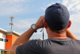 A man peers through binoculars at a telecommunications tower across the road.