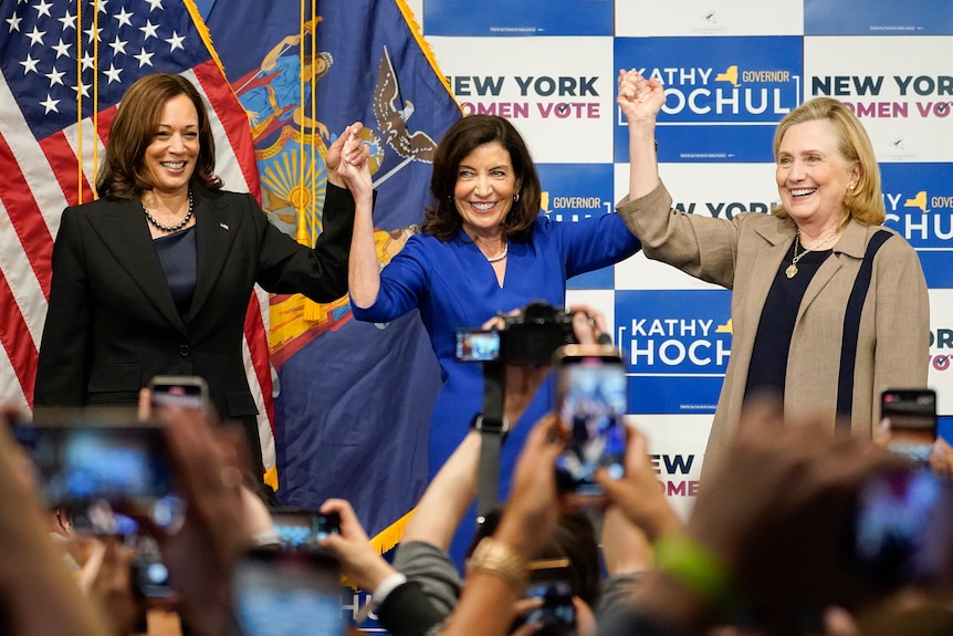 Trei femei care se țin de mână în timpul unei campanii electorale din SUA, în timp ce mulțimea face fotografii. 
