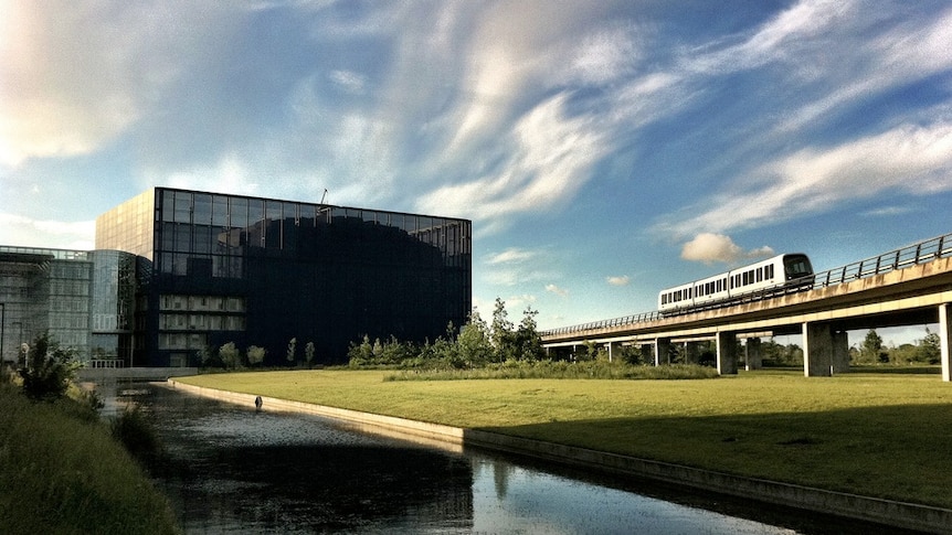 A canal and a large green park lies in front of a black glass office block while an elevated train runs on a track next to it.