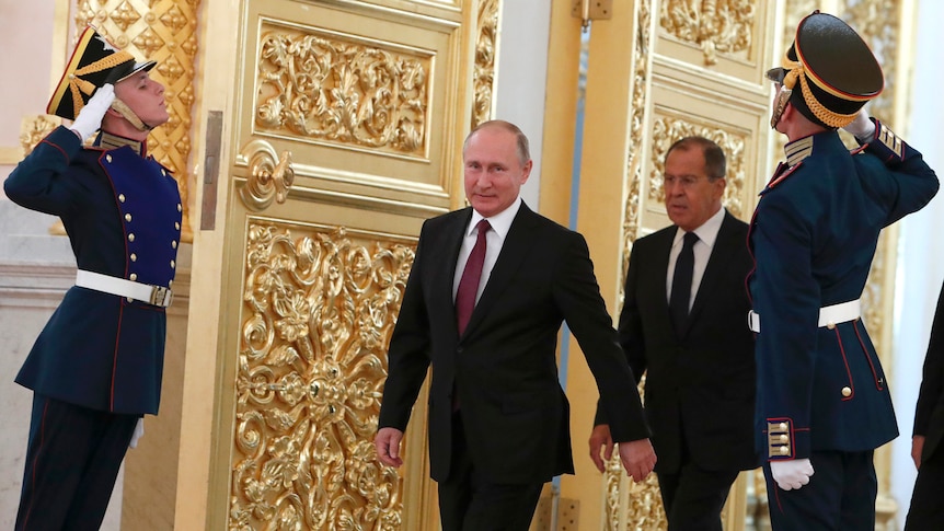 Vladimir Putin and Sergei Lavrov in suits walk past saluting guards in uniform as they walk through large gold doors