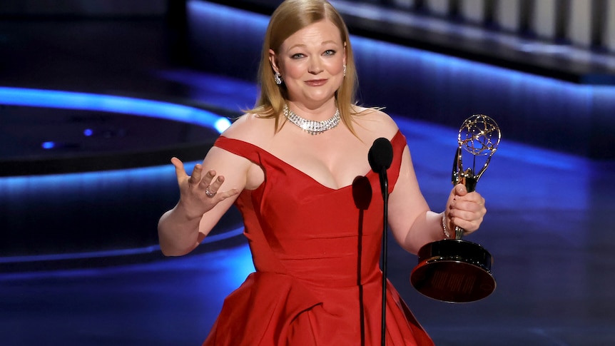 Sarah Snook in a red dress with her arms out holding an Emmy. 