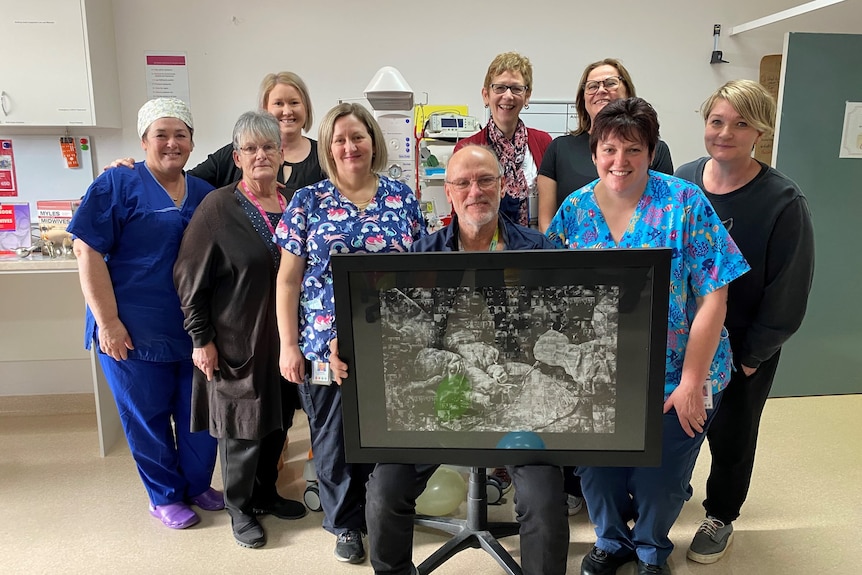 8 hospital staff stand behind a doctor carrying a framed photo of babies.