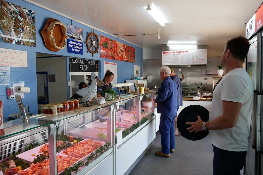 An older gentleman buys fish at a shop counter. A younger man stands with his hat in hand, waiting for his turn.