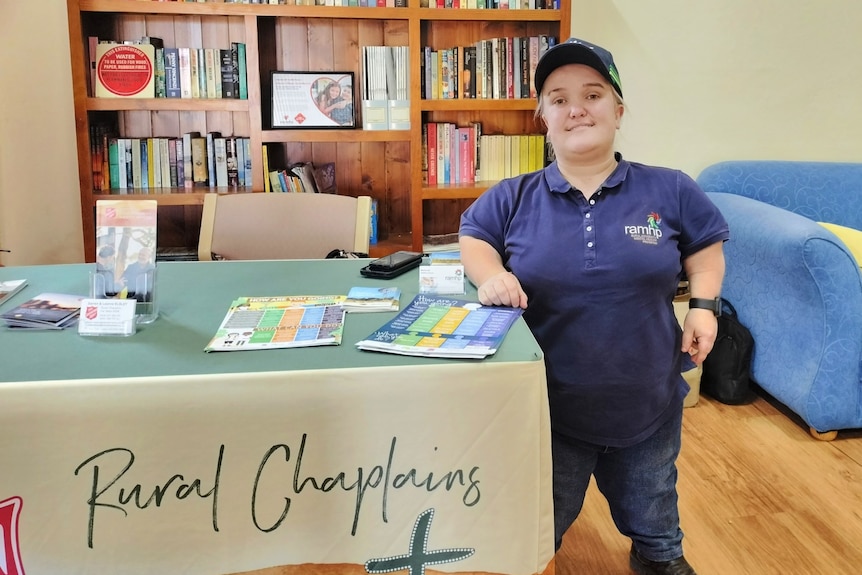 A woman wearing a purple polo shirt stands next to a table with the words Rural Chaplains on it