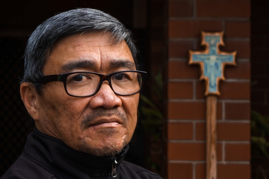 Image of a middle-aged priest with a cross and brick wall in the background