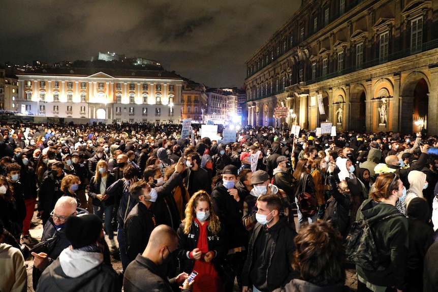 A crowd of people, mostly wearing masks are gathered in a city square at night.