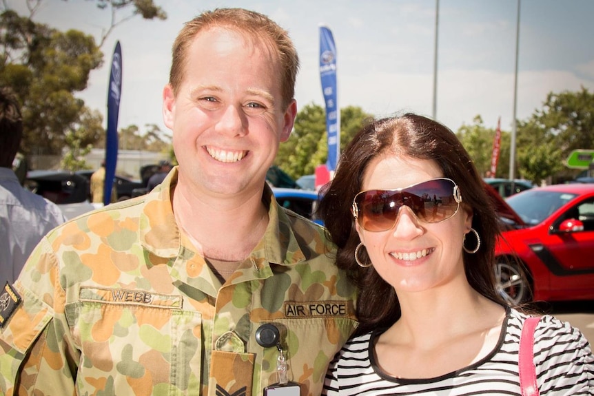 A man in a camouflage army uniform stands smiling for a photo next to a woman and young girl wearing a bucket hat.