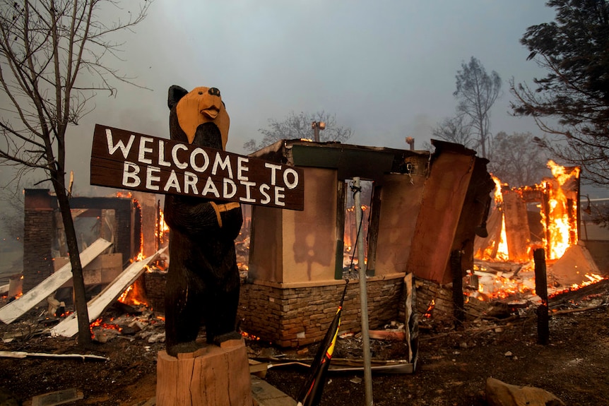 A sign at a campsite with a burning building behind it as wildfire rips through township.