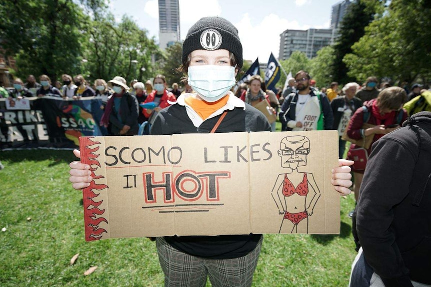 A young person wearing a face mask holds a placard reading "Scomo likes it hot" as they stand in front of demonstrators.
