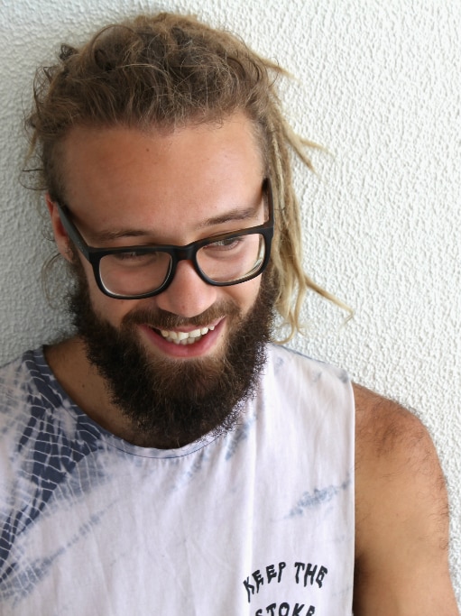 man with dreadlocks, beard and glasses standing against a wall, looking down and smiling