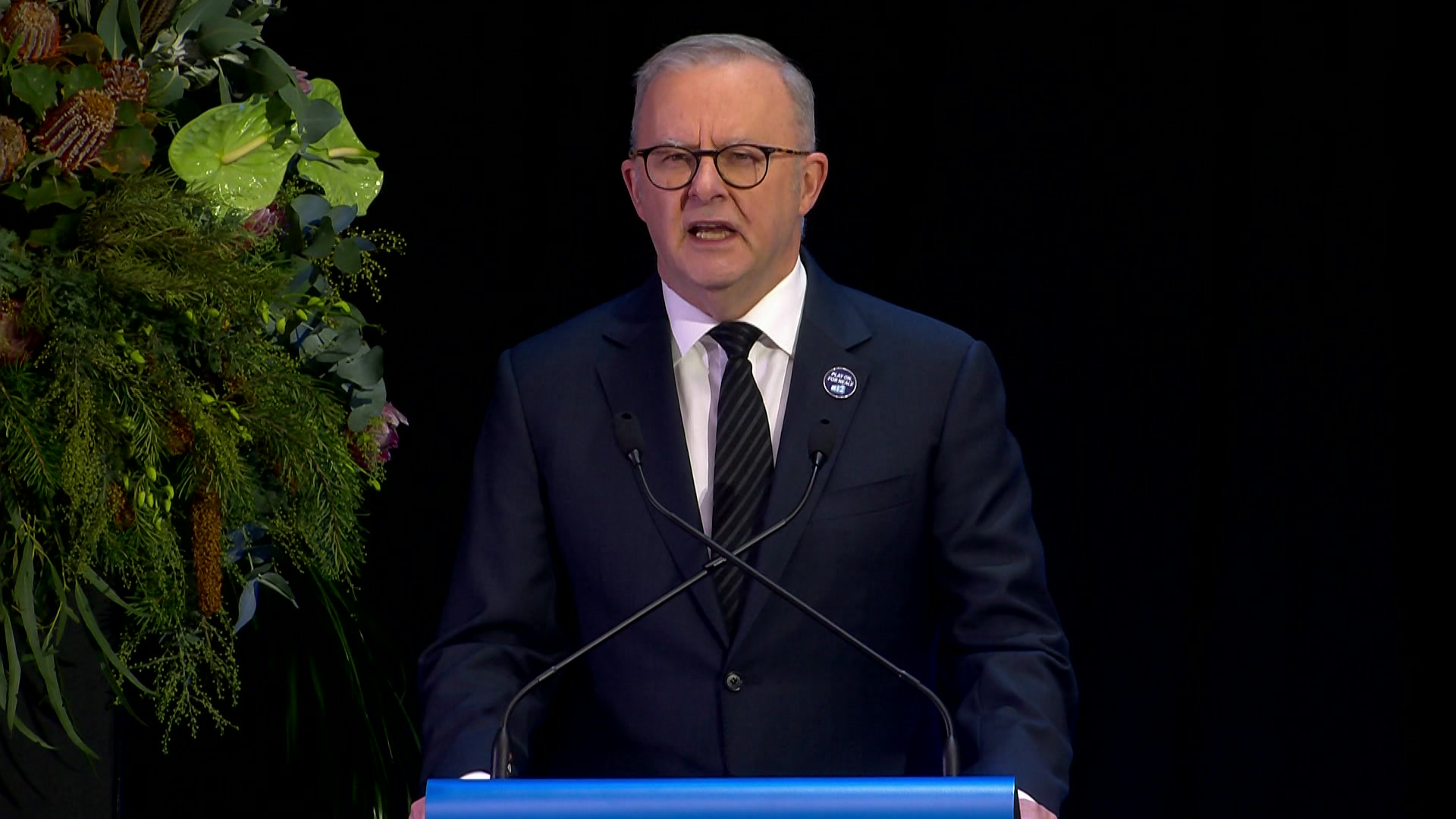 A man with grey hair, dark rimmed glasses in a dark suit and tie talks into a microphone at a lecturn.