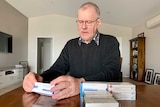 A man wearing a collared shirt, jumper and glasses sits at a kitchen table reading a medication box, surrounded by other boxes.