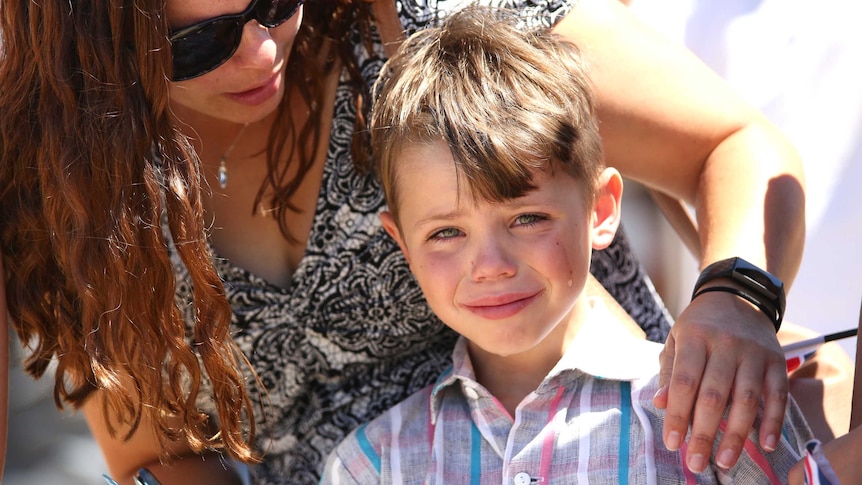 A boy dressed in a collared shirt smiles through a cry as he's comforted by an adult