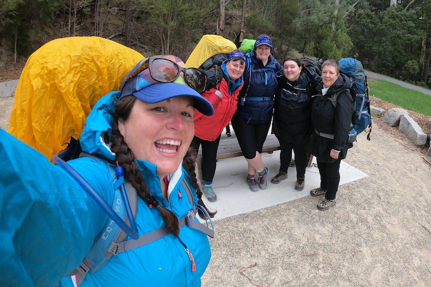 Five women stand smiling at the camera, they all carry packs and are dressed in wet-weather gear.