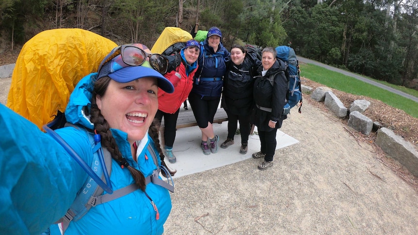 Five women stand smiling at the camera, they all carry packs and are dressed in wet-weather gear.