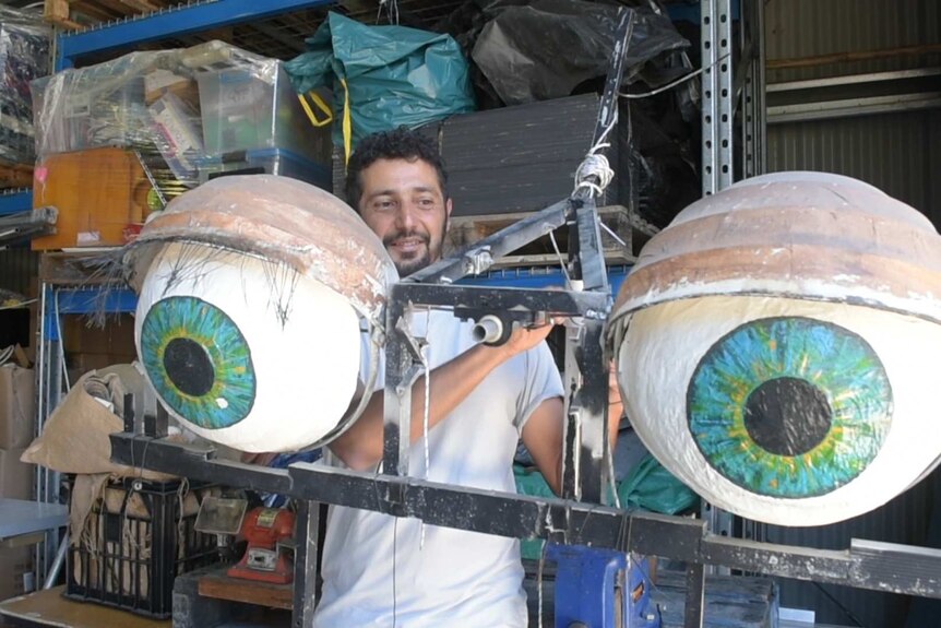 A man with dark curly hair and a beard standing behind two giant papier mache eyeballs.