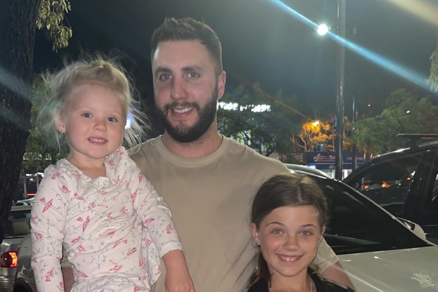 A smiling young bearded man with dark hair cradling a little girl. He has his arm around another young girl.