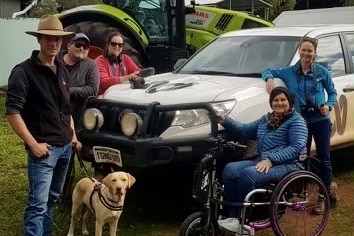 Group of people standing next to ABC 4WD, one woman in a wheelchair and a farmer with a guide dog.