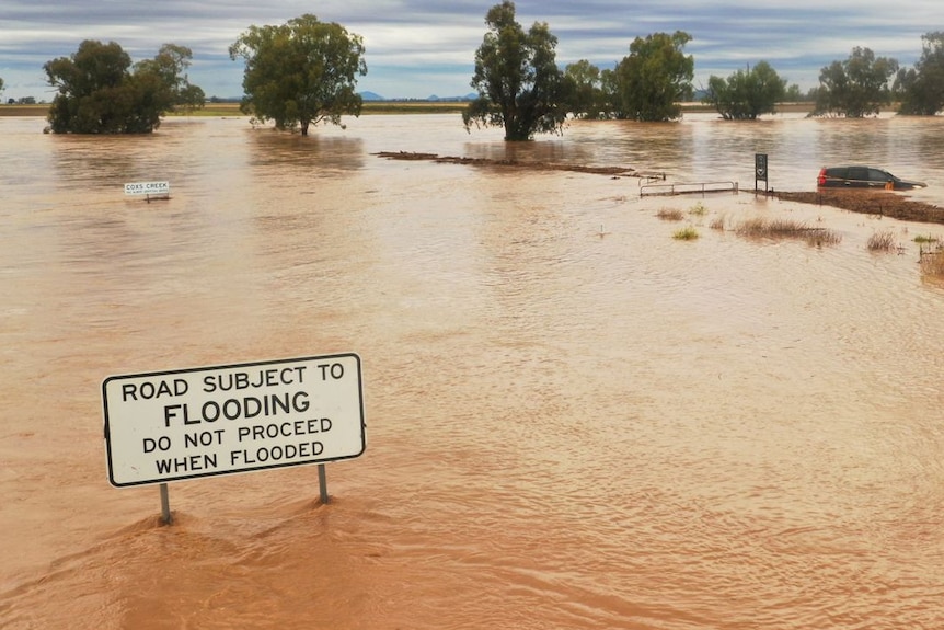 A sign telling cars not to proceed in floodwaters, with a car flooded in behind it.
