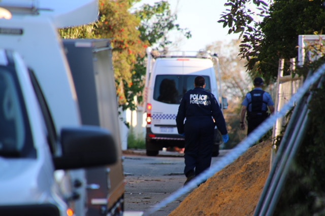 A police forensic officer in a blue jumpsuit walks behind a police vehicle on a Perth street near a pile of sand.