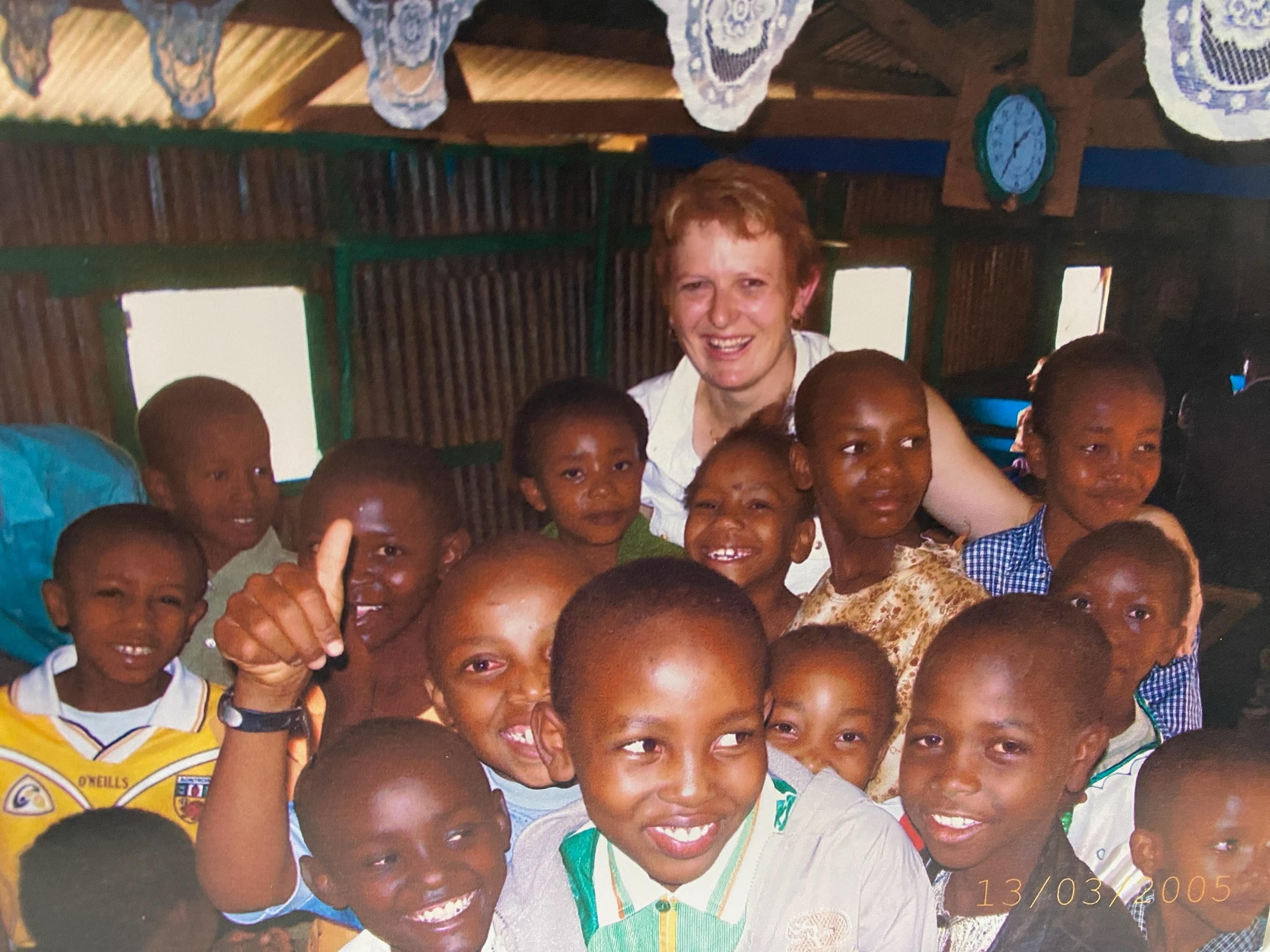 Una mujer está detrás de un gran grupo de niños, todos vestidos con camisas de diferentes colores, sonriendo. 