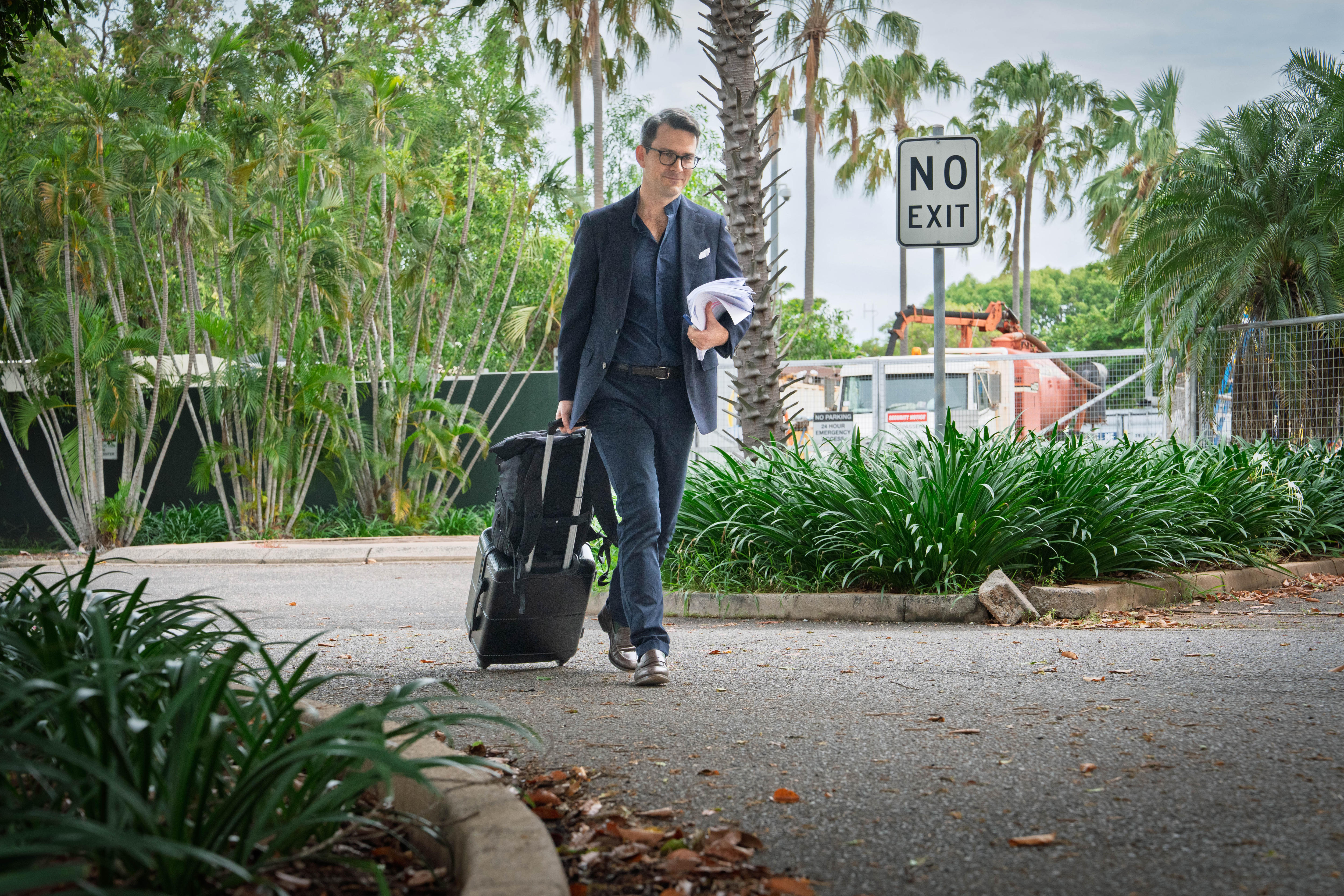 Um homem branco de terno azul escuro: paletó, calça, camisa, todos da mesma cor. Andando com mala preta, segurando documentos em papel.