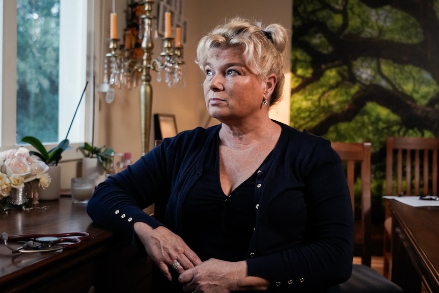 Woman with blonde hair tied up, wearing a navy sweater sitting at a desk and looking out a window.