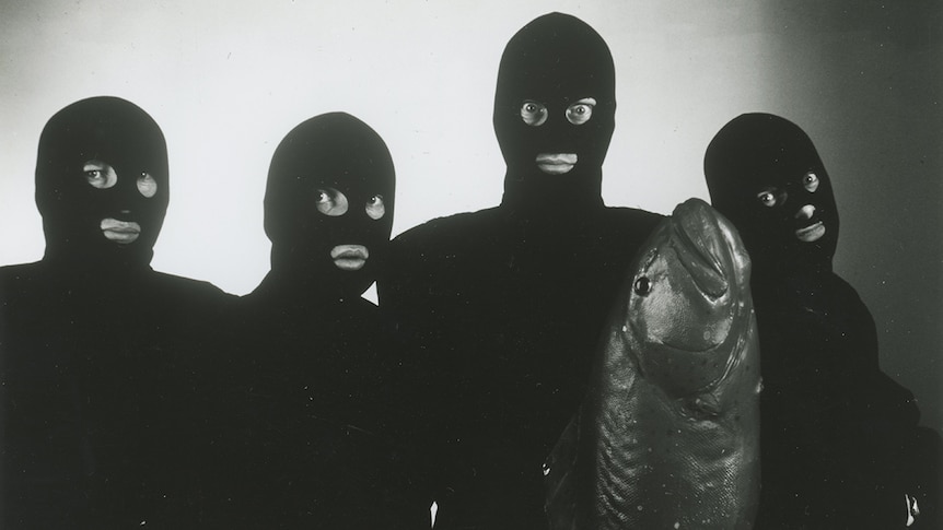Black and white photo of four men wearing black balaclavas and body suits, holding a large fish