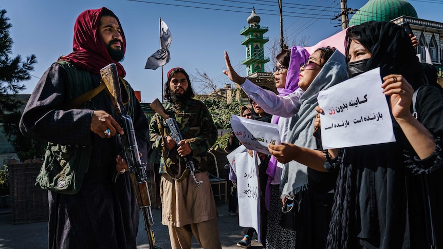 Group of women, heads draped in scarves, hold placards in front of a Taliban soldier holding a gun.