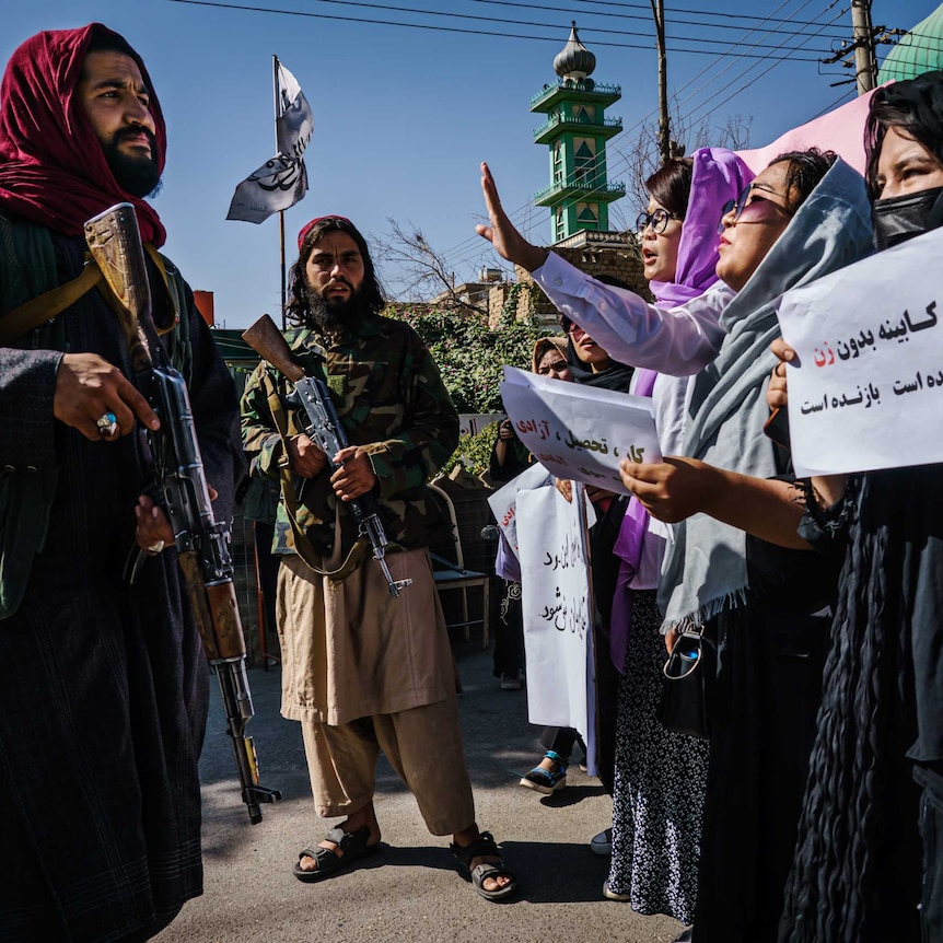 Group of women, heads draped in scarves, hold placards in front of a Taliban soldier holding a gun.