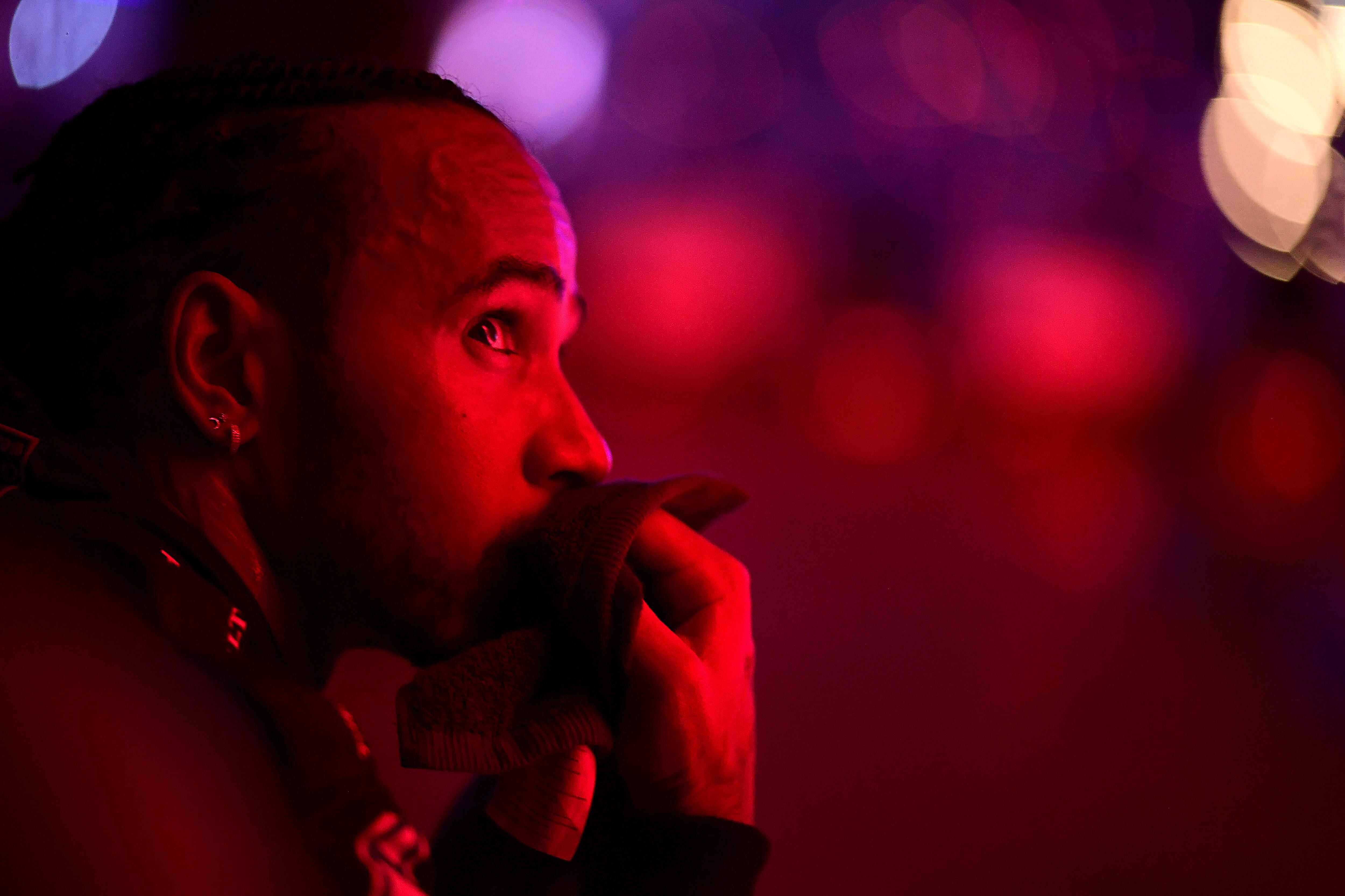 Lewis Hamiton, sitting down, holding a towel to his face, looks upward with a red light shining on him, at night after a race