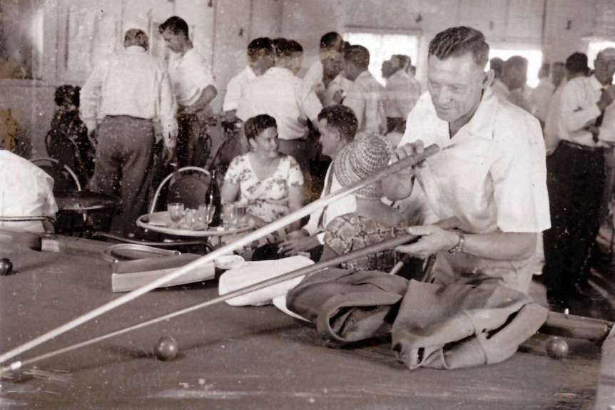 A black and white photo of people socialising a man plays pool in the foreground