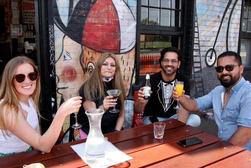 Two women and two men sit at an outdoor table beside a graffiti wall and hold up their drinks while smiling at the camera.