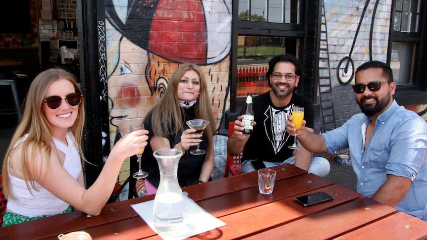 Two women and two men sit at an outdoor table beside a graffiti wall and hold up their drinks while smiling at the camera.