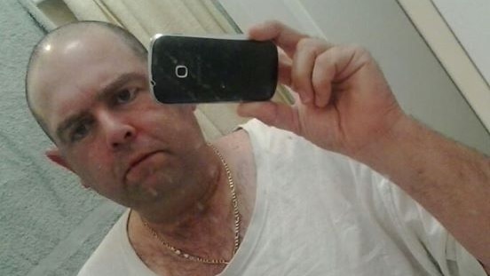 A man in a white t-shirt takes a selfie in a mirror with a black mobile phone.