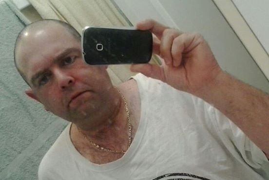 A man in a white t-shirt takes a selfie in a mirror with a black mobile phone.