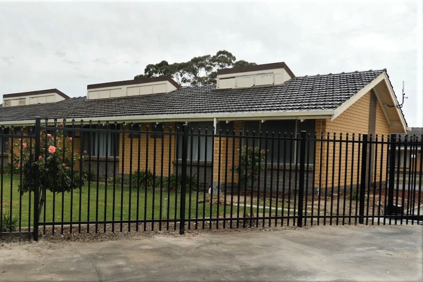 The exterior of a motel. Rose bushes planted behind a black metal fence, the building has a bright green lawn in front