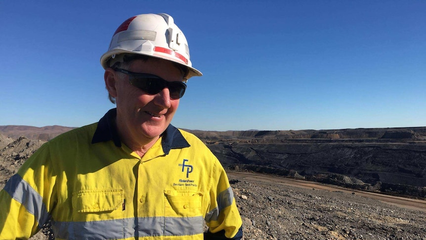 A man in fluoro safety equipment and sunglasses stands atop a coal mine. Huge dark mounds of dirt are in the background.