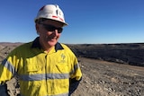 A man in fluoro safety equipment and sunglasses stands atop a coal mine. Huge dark mounds of dirt are in the background.