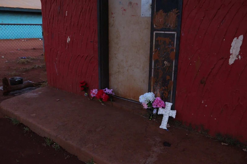 A cross and some floral tributes to Kumanjayi Walker lean against the deep red wall of a house in Yuendumu.