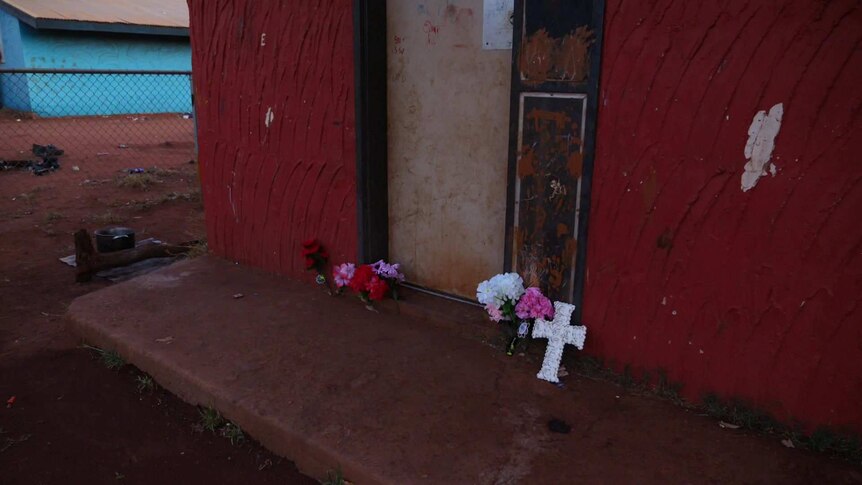 A cross and some floral tributes to Kumanjayi Walker lean against the deep red wall of a house in Yuendumu.