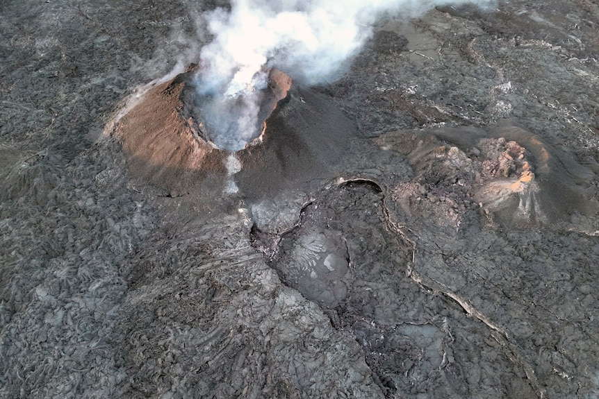 A white cloud of smoke billows from a volcano that is photographed from the top-down by an aeroplane against a black landscape