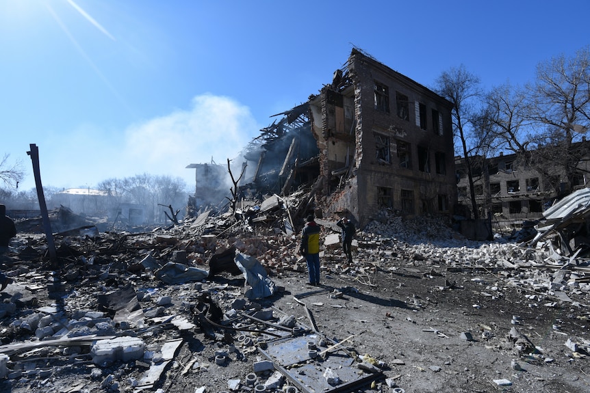 People stand in front of a destroyed shoe factory in the aftermath of a missile attack.