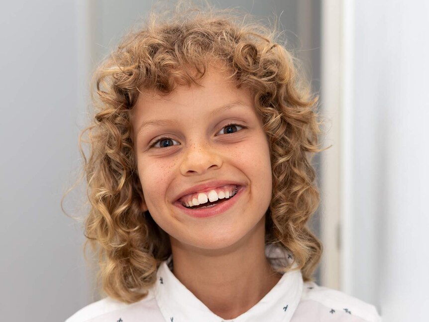 A boy with blond curly hair in a white shirt with a big smile