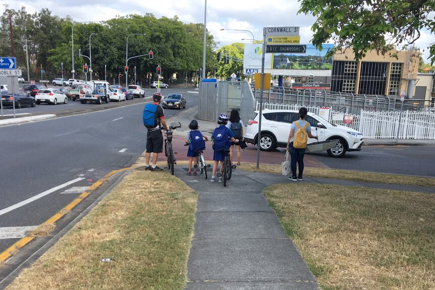 A parent with three schoolkids on bikes wait for a safe moment to cross a busy road